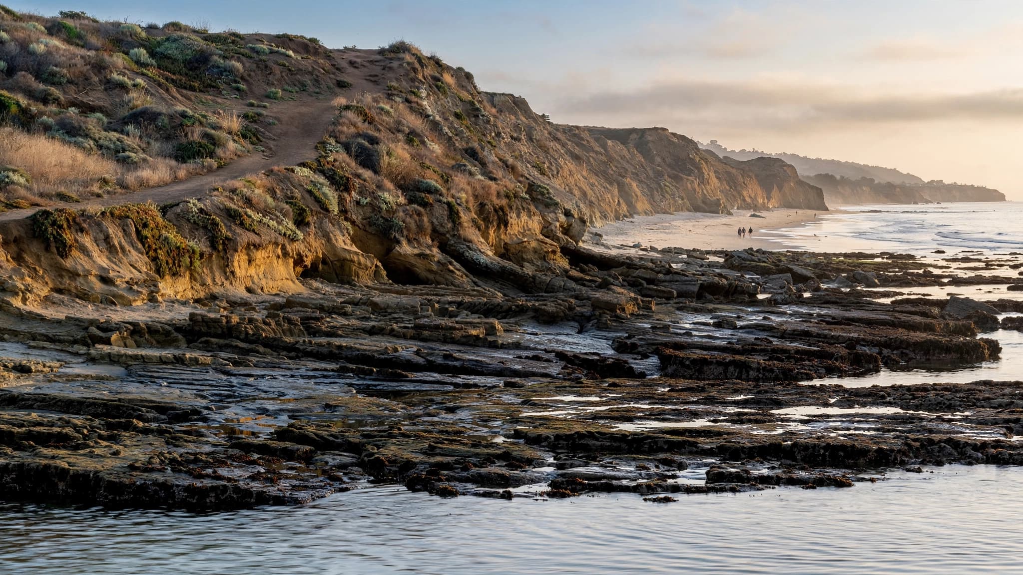 Rocky Malibu coastline at El Matador State Beach at low tide in late-afternoon light, with a dirt bluff path above and calm w