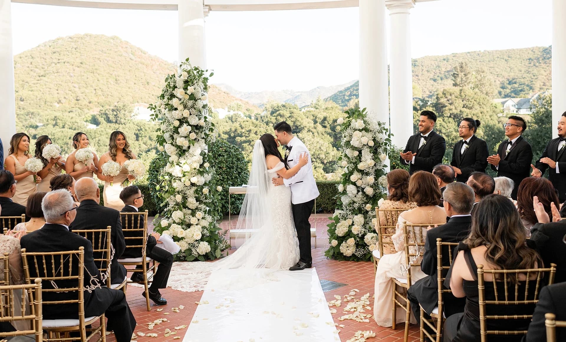 Newlywed couple sharing a kiss at their outdoor wedding ceremony altar in front of guests.